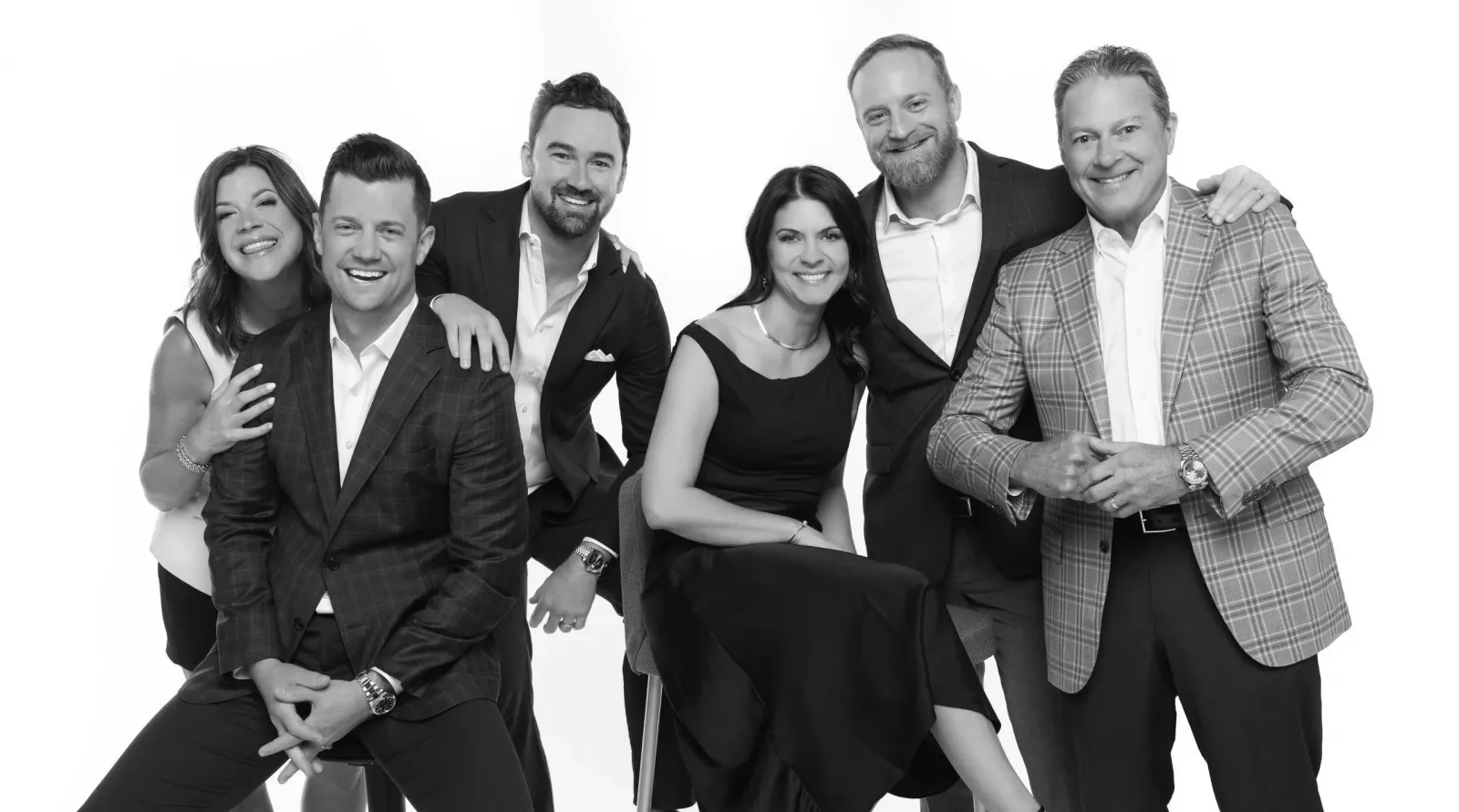Six adults in professional business attire stand together, smiling, against a plain white background in a black-and-white photo.
