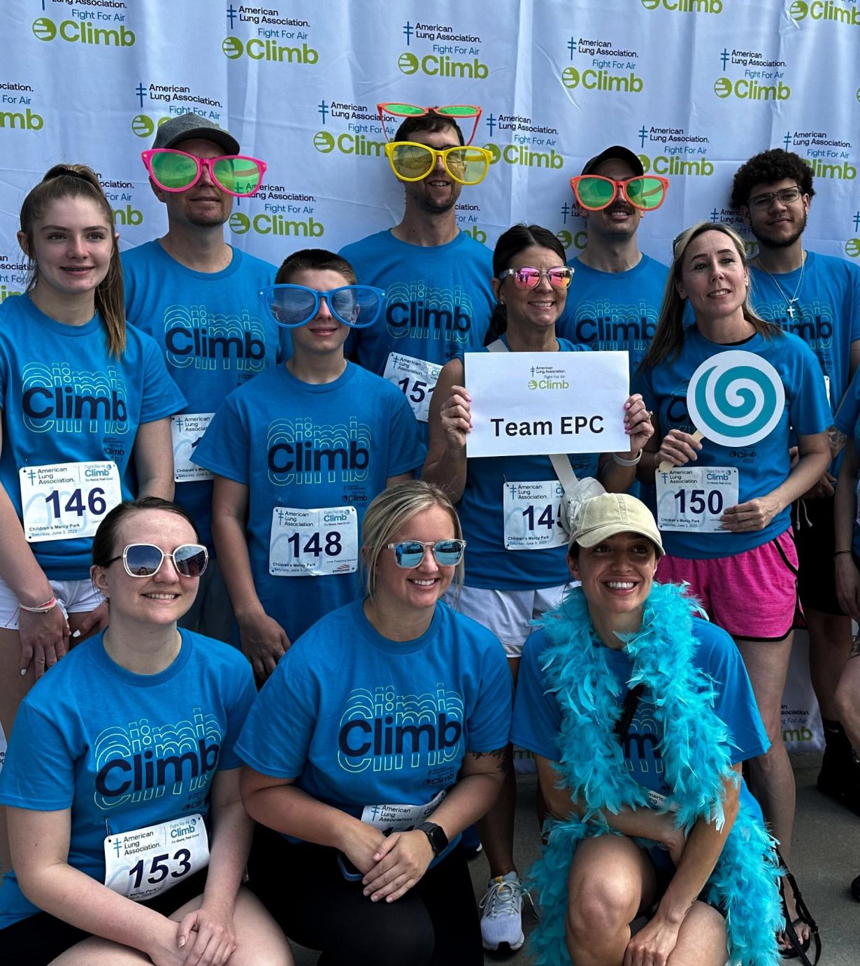 A group in blue "Climb" shirts poses with race bibs and fun accessories, holding a "Team EPC" sign.