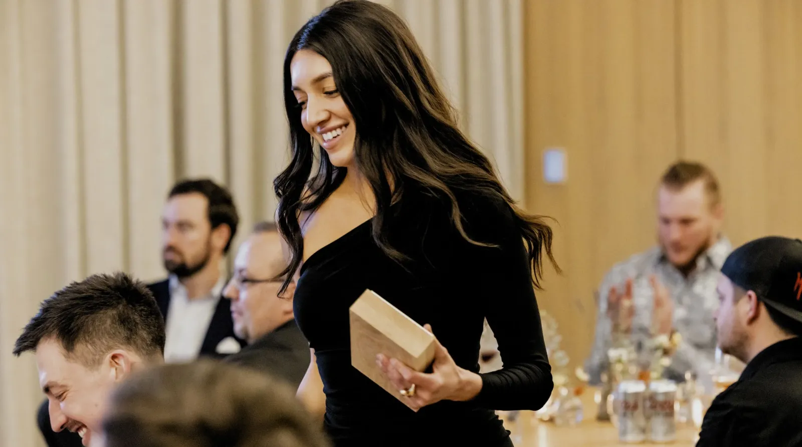A woman in a black dress, holding a wooden box, stands and smiles in an event room with seated people at tables.