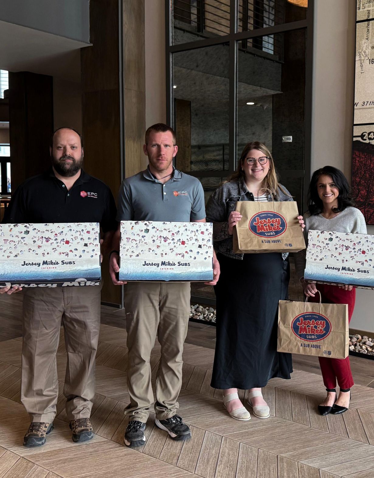 Four people standing indoors holding two large decorated boxes and two Jimmy John’s bags, smiling at the camera.