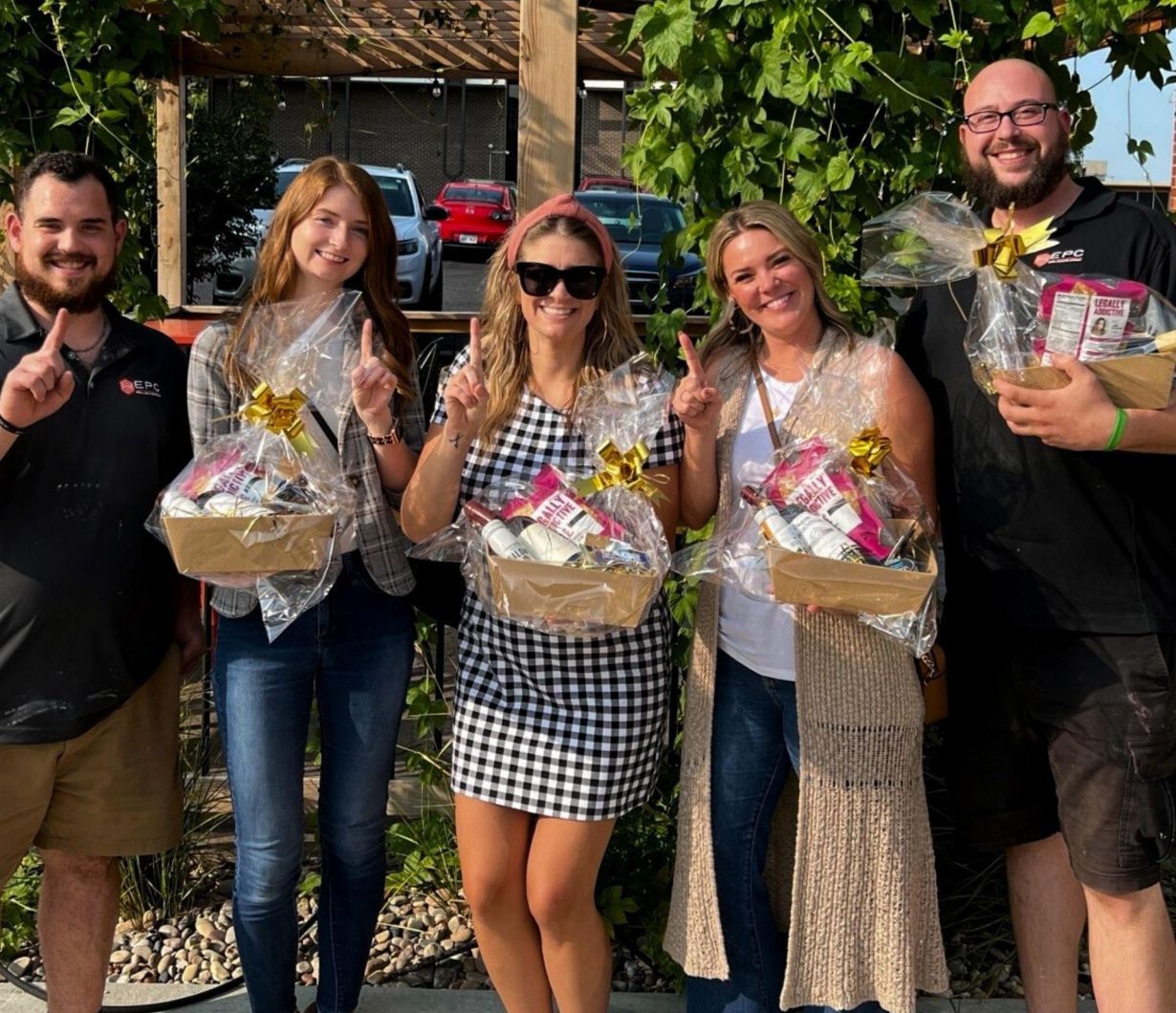 Five smiling people outdoors holding gift baskets and making peace signs, with greenery in the background.