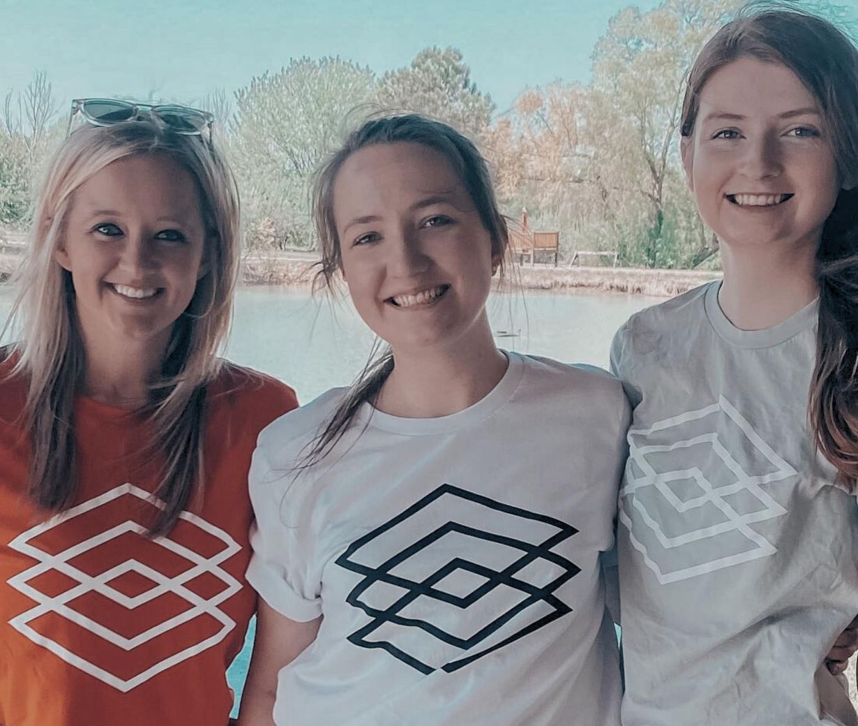 Three women smiling outdoors, wearing geometric-patterned t-shirts, with trees and water in the background.